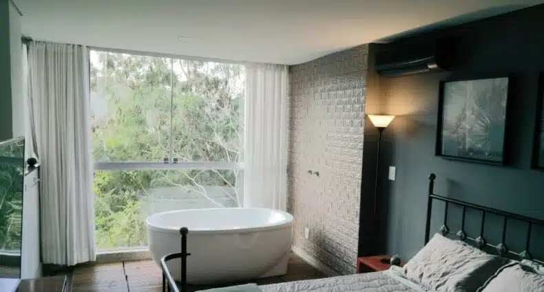 Foto do Loft Espaço Vila da Serra. O ambiente recebe bastante iluminação. A cama de casal pode ser vista no canto inferior direito da foto, e atrás há uma parede verde. Seguindo ao fundo do quarto, encostado na janela, há uma banheira de hidromassagem, com uma vista da serra da Mantiqueira, para representar pousadas em Nova Lima.