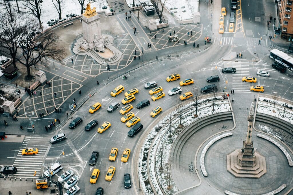 Foto vista de cima de uma rotatória em Nova York, no bairro Columbus Circle, com vários carros e táxis amarelos contornando o monumento de pedra no centro da rotatória. É um dia cinzento e há muita neve ao redor, além de pessoas caminhando nas calçadas. Essa é uma das fotos para representar o post de aluguel de carro em Nova York.