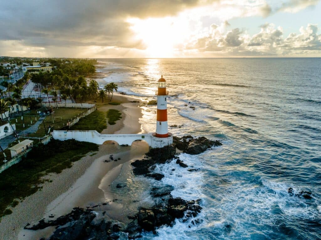 Um farol branco e vermelho em uma praia de areia clara, pedras e mar azul claro no litoral de Salvador na Bahia, para representar seguro viagem nacional