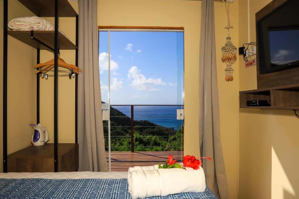 Quarto do Flat Vista Mar Noronha com cama, TV, armário aberto e uma janela aberta mostrando uma varanda com vista para o mar e o céu nublado.