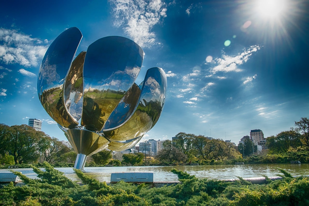 Escultura de flor de metal refletindo vegetação e um espelho d'água. O céu acima é azul com poucas nuvens. - Foto: 3345557 via Pixabay