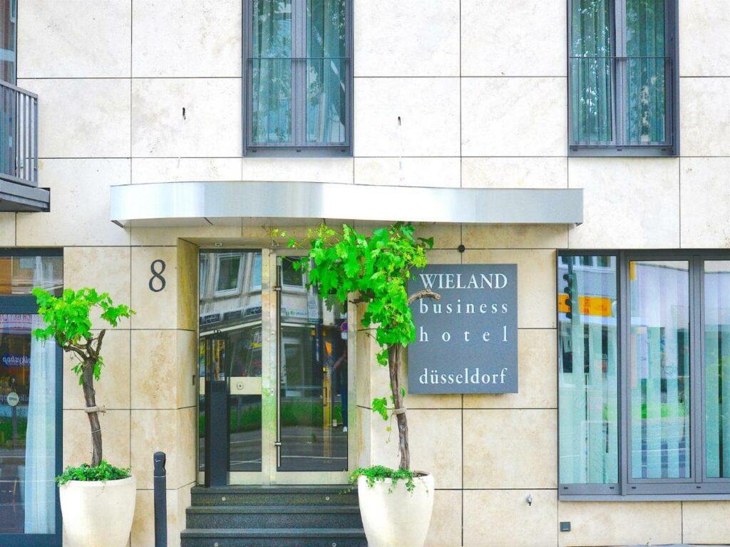 Frente do Business Wieland Hotel com porta no centro da imagem com dois vasos com árvores.