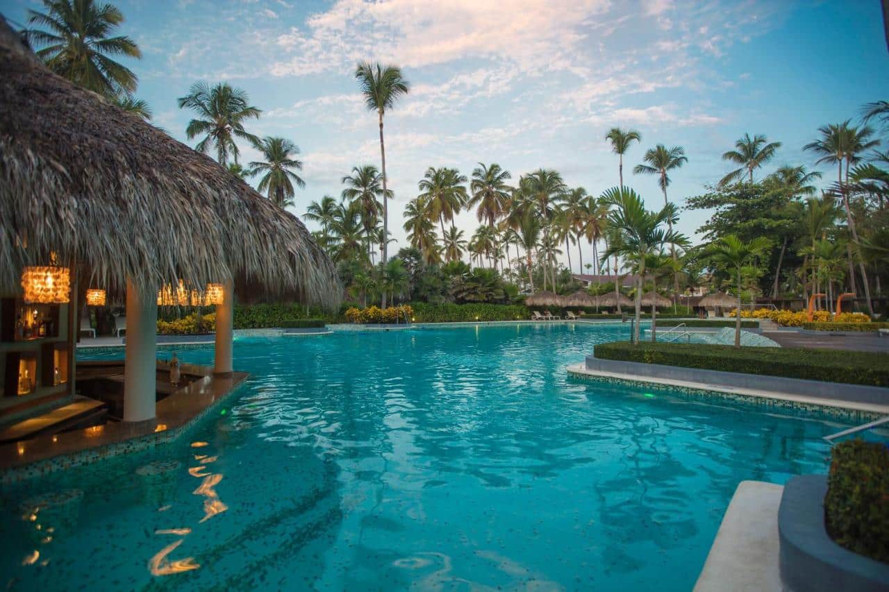 Piscina do Grand Palladium Bavaro Suites Resort & Spa cercada por palmeiras e com uma estrutura de palha no canto esquerdo.