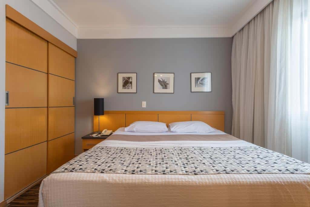 Quarto do Green Place Ibirapuera. Uma cama de casa no meio, do lado esquerdo uma cômoda com uma luminária, no lado direito a janela do quarto com cortinas fechadas, do lado esquerdo o guarda-roupa. Foto para ilustrar post sobre hotéis perto do Parque Ibirapuera.