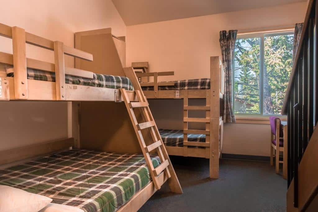 Quarto do HI Banff Alpine Centre - Hostel com duas beliches de madeira com travesseiros e cobertas xadrez. Ao lado de um dos beliches tem uma janela de vidro com vista para a árvores, ilustrando post Hotéis em Banff.
