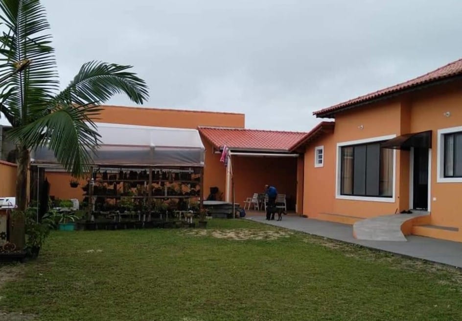 Área externa da Hospedagem Familiar Yamane. Fundo da hospedagem no lado direito, no lado esquerdo um jardim com várias plantas no fundo. No eio, uma pessoa com um cachorro.