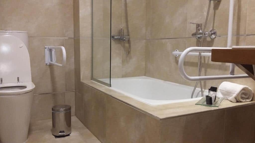 Banheiro com acessibilidade da Hostería Huenú. Um vaso sanitário do lado esquerdo, com uma barra e um lixo. Do lado direito, a banheira com o bxz e o chuveiro. Uma barra de apoio para entrar, e duas barras no box.