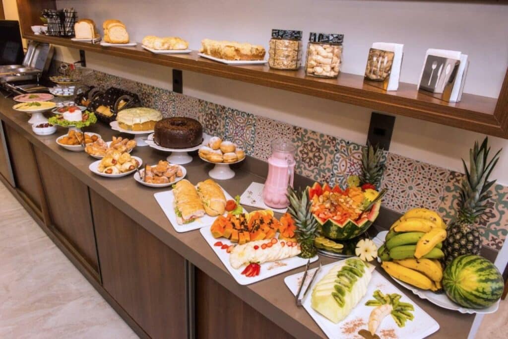 Café da manhã do Hotel Balneário do Parque. Em uma bancada e uma prateleira estão vários tipos de frutas, pães, bolos, doces e salgados e outras coisas para comer.