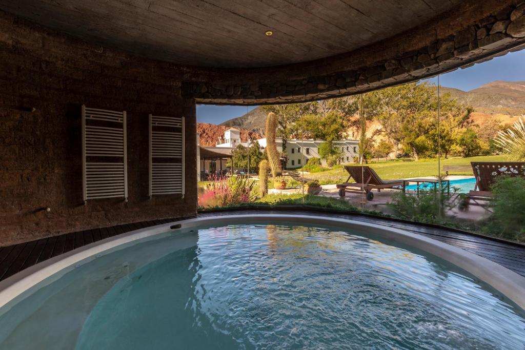 Espaço fechado com banheira de hidromassagem do Hotel El Manantial del Silencio. Uma parede de vidro do lado direito com vista para a piscina, cadeiras de sol, cactos e montanhas.