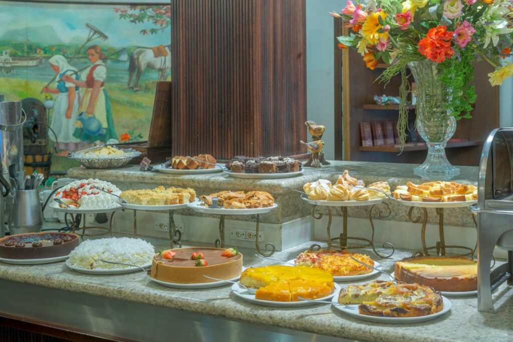 Café da manhã do Hotel Glória. Diversos tipos de bolos doces, tortas, pães e salgadoes estão em um balcão.