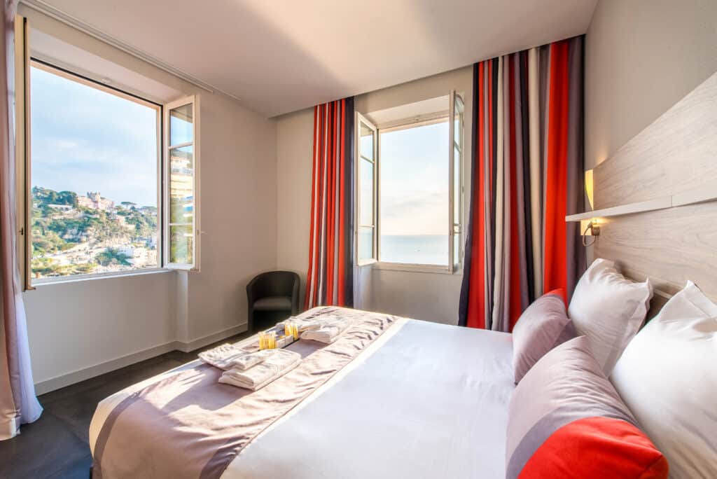 Quarto do Hotel Le Saint Paul. A cama fica encostada na parede direita do quarto e há duas janelas grandes com vista para o mar, uma em frente e a outra no lado da cama. Imagem para ilustrar o post hotéis em Nice.