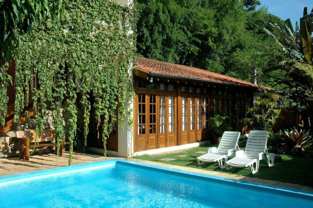 Área aberta da piscina do Hotel Pousada São Rafael. A piscina está rodeada por plantas e duas espreguiçadeiras. Ao fundo está a própria pousada.