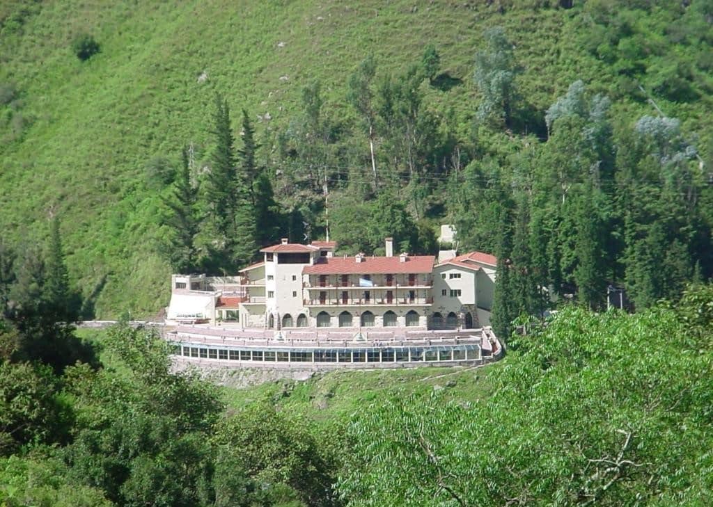 Vista do Hotel Spa Termas de Reyes. O prédio grande do hotel no meio, atrás uma montanha e ao redor várias árvores.