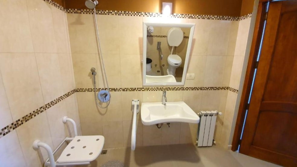 Banheiro com acessibilidade do Hotel Vitalia. Um acento no lado esquerdo bom barras e o chuveiro em cima, no meio, uma pia rebaixada com espelho e barras, no lado direito a porta do banheiro.