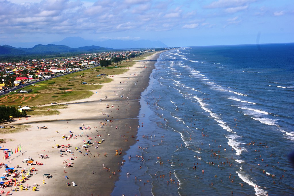 Foto da praia de Ilha Comprida. Do lado direito o mar com ondas calmas. Do lado esquerdo a areia e a cidade atrás. Na areia e no mar várias pessoas.