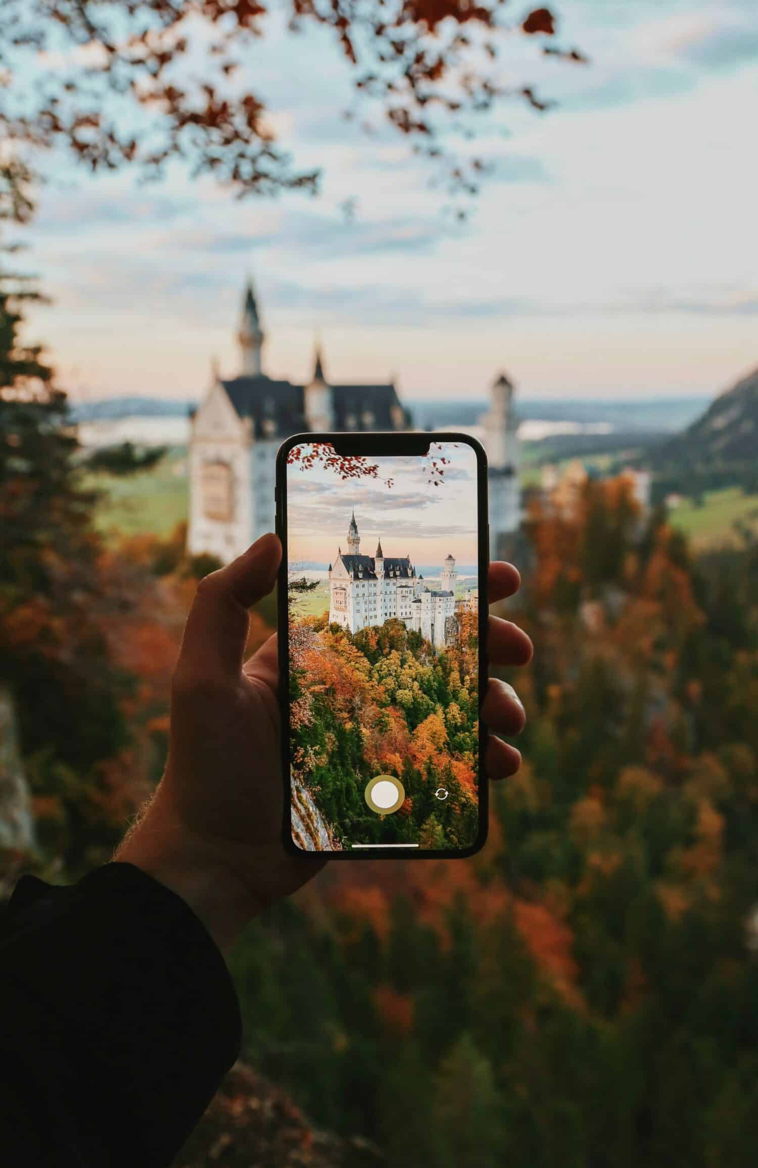 mão branca adulta segurando um celular para tirar foto do Castelo de Neuschwanstein ao fundo, com várias árvores e vegetações em tons marrom e laranja