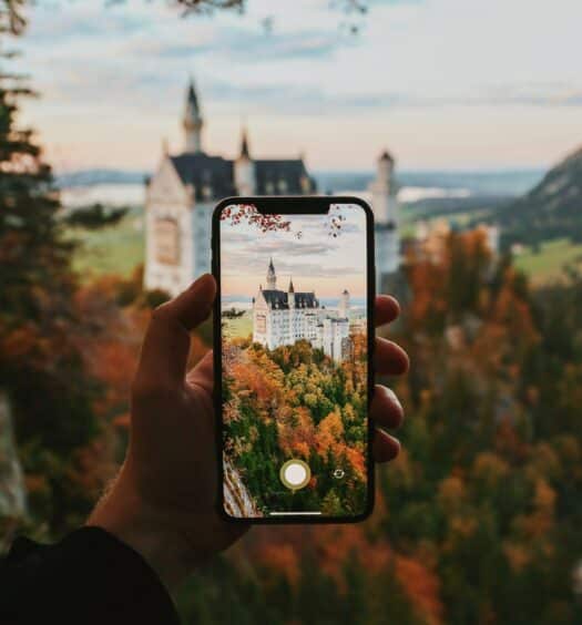 mão branca adulta segurando um celular para tirar foto do Castelo de Neuschwanstein ao fundo, com várias árvores e vegetações em tons marrom e laranja