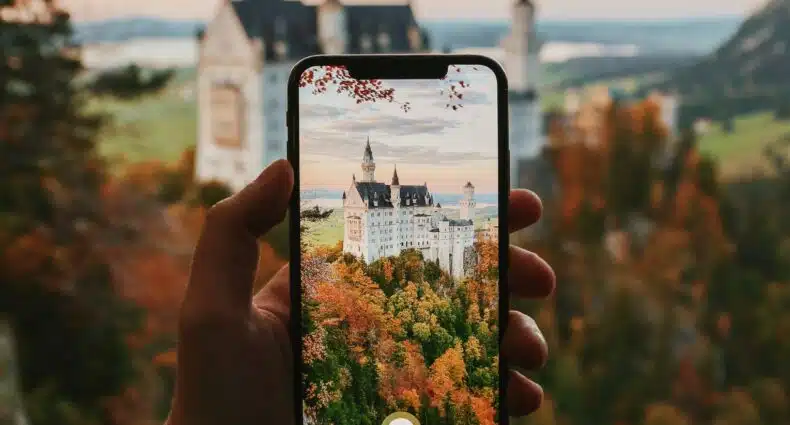 mão branca adulta segurando um celular para tirar foto do Castelo de Neuschwanstein ao fundo, com várias árvores e vegetações em tons marrom e laranja