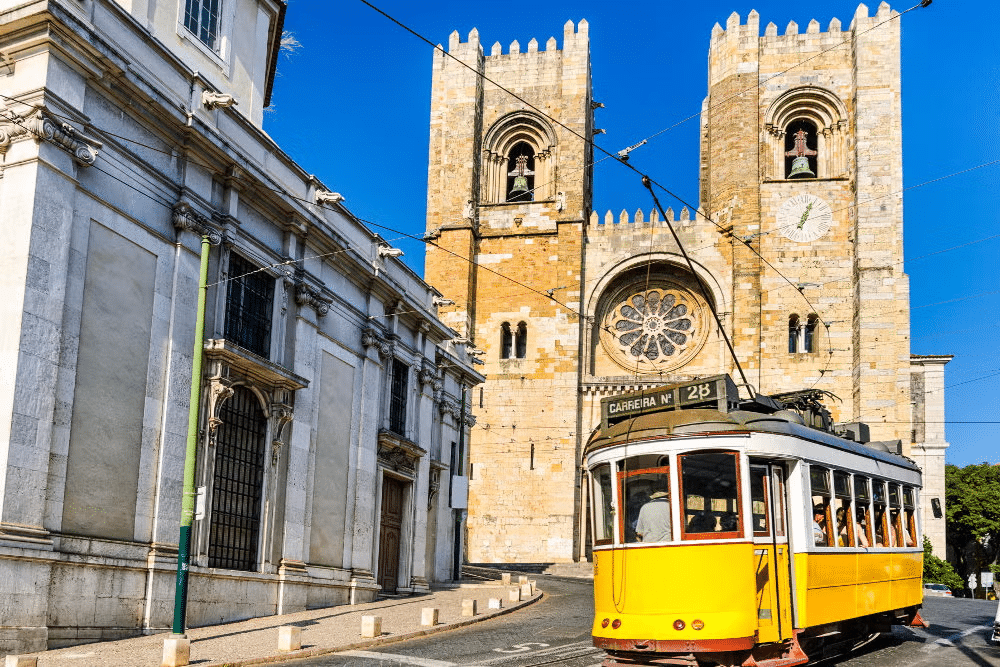 Vista da Praça do Rossio, em Lisboa, com uma igreja imponente com vitrais e torres com sinos grandes, além de um bondinho elétrico na frente e um prédio com fachada histórica