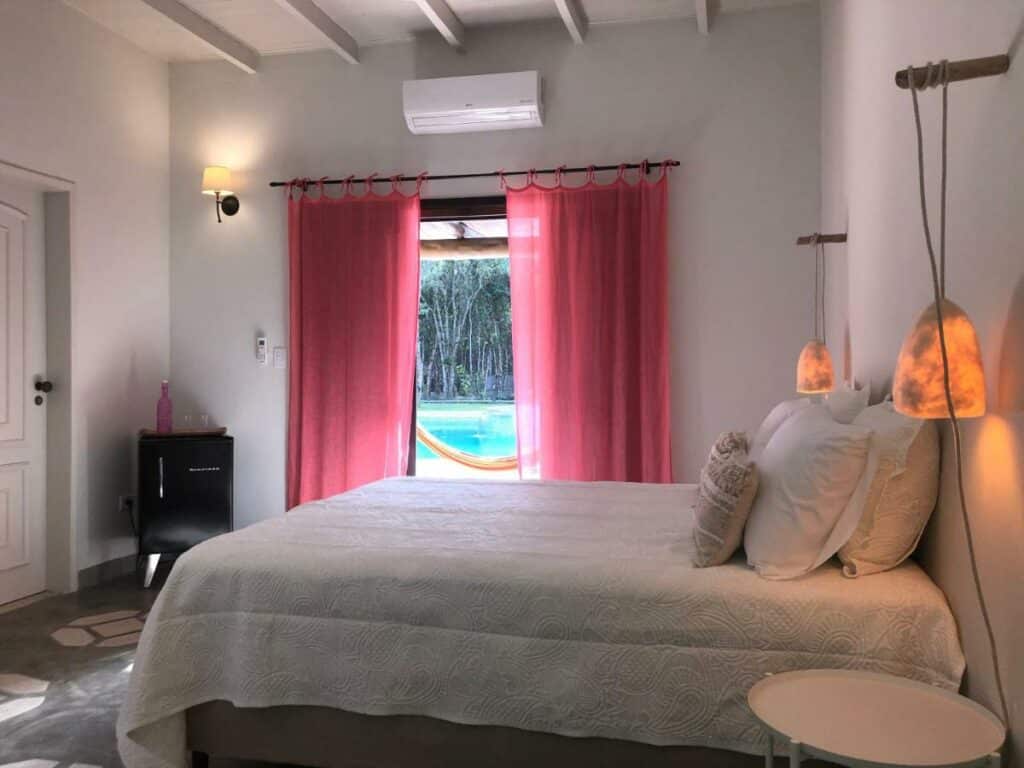 Quarto da pousada Jardim Boêmio. Uma cama de casal está no meio, com duas luminárias penduradas em seus dois lados. Em frente há uma porta e um mini frigobar. Ao lado da cama está uma porta com cortinas, que leva para a área da piscina.