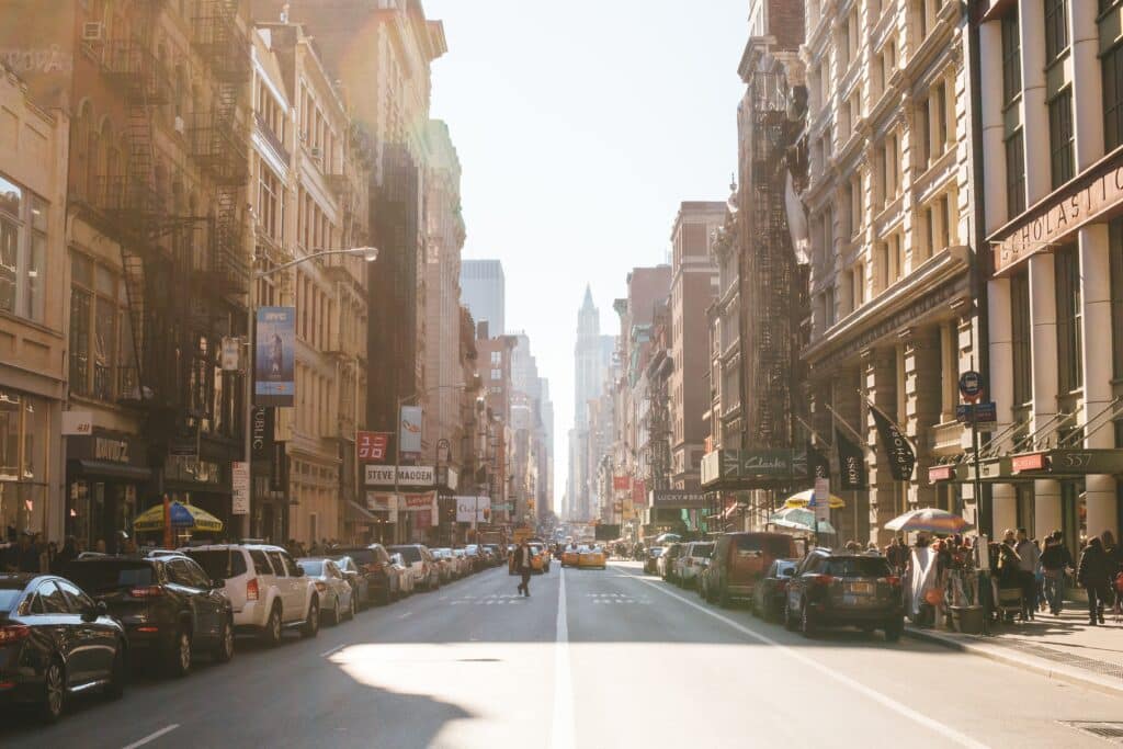.Ruas do Brooklyn em Nova York para representar o post aluguel de carro em Nova York. A foto mostra uma avenida no centro com vários prédios antigos margeando as ruas, além de carros e pessoas passeando. O dia está claro e ensolarado.