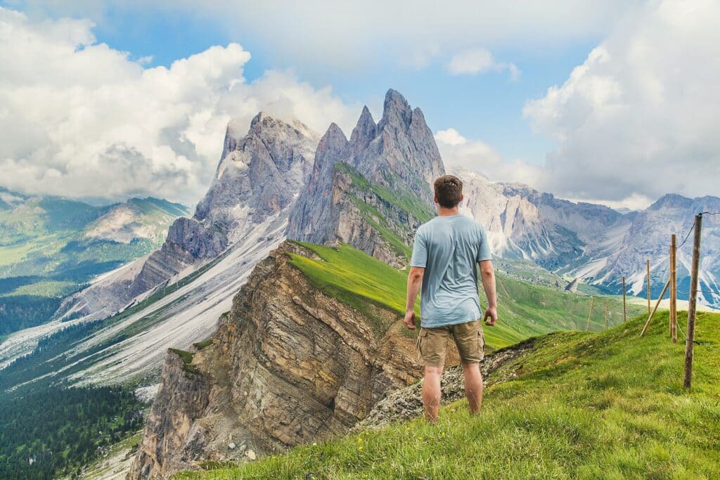Um homem de bermuda e camiseta em cima de uma planície irregular olhando para diversas montanhas cobertas por grama, para representar seguro viagem internacional