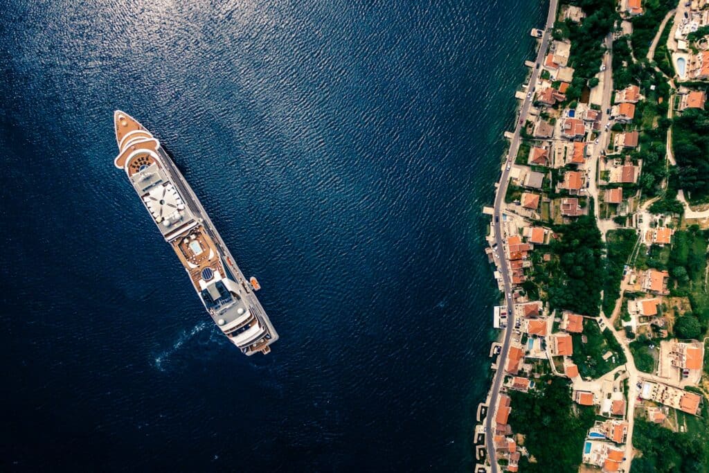 Foto aérea de um navio passando perto da costa de uma cidade. O mar tem águas escuras e o navio segue em direção ao canto superior esquerdo. Pode-se ver tetos de casinhas cercadas por vegetação ao lado direito. - Foto: Polina Rytova via Unsplash