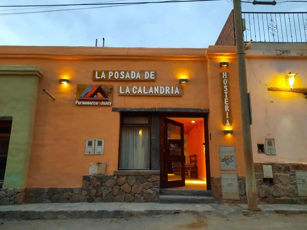 Frente da La Posada de la Calandria. A placa com o nome da hospedagem, dois degraus e a porta de vidro do local. No lado direito um poste.