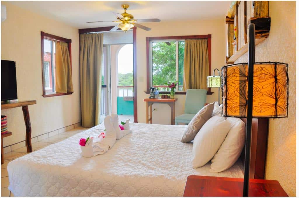 Foto de um quarto no Lamanai Hotel & Marina. Vemos uma cama em um ângulo lateral. Assim, percebemos uma mesa de cabeceira com abajur em cima ao lado direito da cama. E na frente dela. há uma mesa de madeira com TV, e ao lado dessa TV, há uma janela. Já no outro lado da cama, mais ao fundo da quarto, vemos uma cadeira ao lado de outra mesa de madeira que possui amenidades em cima, e atrás há mais uma janela. A porta do quarto está entre as duas janelas descritas.