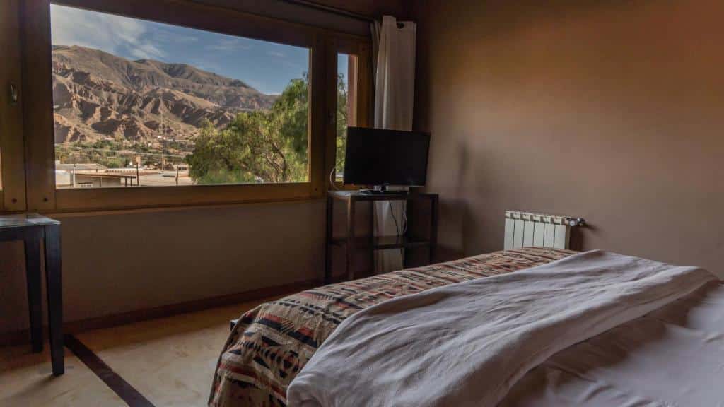 Quarto da Las Marías Hotel Boutique. Uma cama de casal do lado direito, de frente uma mesa com uma televisão e a janela do quarto com vista para as montanhas.