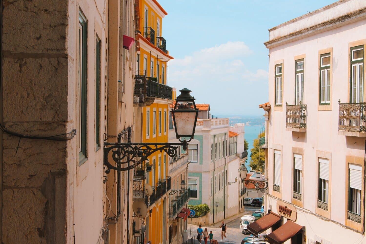 Rua inclinada em Lisboa, com prédios coloridos em cada lado da rua, rio ao fundo, e uma lanterna externa em primeiro plano, para ilustrar o post de destinos mais visitados da Europa