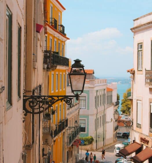 Rua inclinada em Lisboa, com prédios coloridos em cada lado da rua, rio ao fundo, e uma lanterna externa em primeiro plano, para ilustrar o post de destinos mais visitados da Europa
