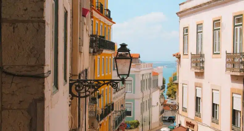 Rua inclinada em Lisboa, com prédios coloridos em cada lado da rua, rio ao fundo, e uma lanterna externa em primeiro plano, para ilustrar o post de destinos mais visitados da Europa