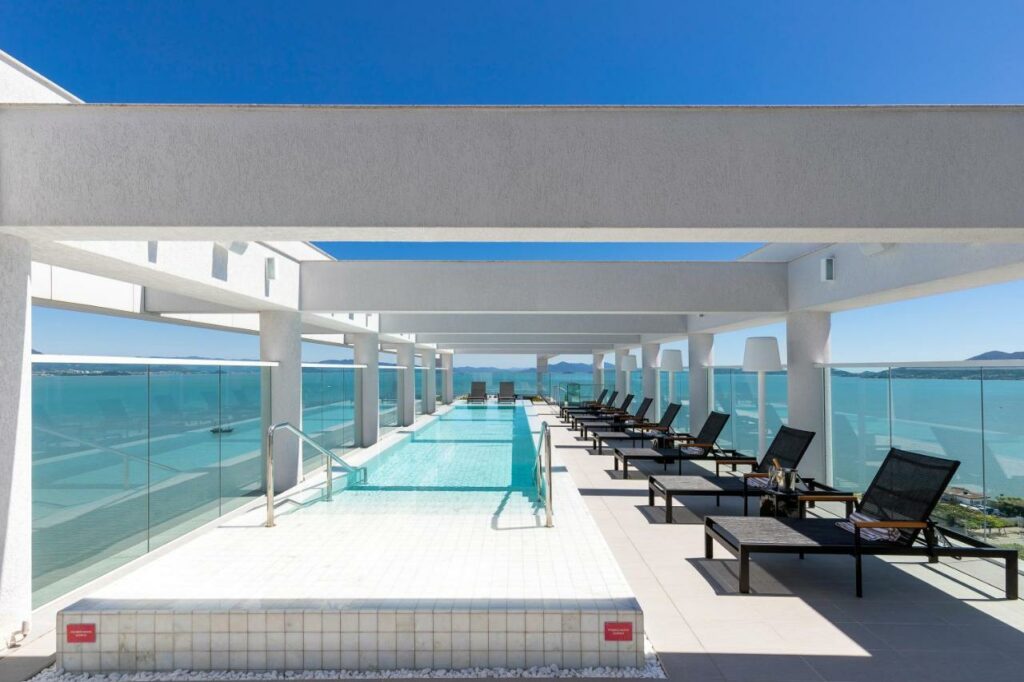 Área da piscina do LK Design Hotel Florianópolis.. A piscina está do lado esquerdo, tem formato retangular, é fina e bem comprida. Do lado direito estão várias espreguiçadeiras enfileiradas. A área é a céu aberto e tem vista para o mar.