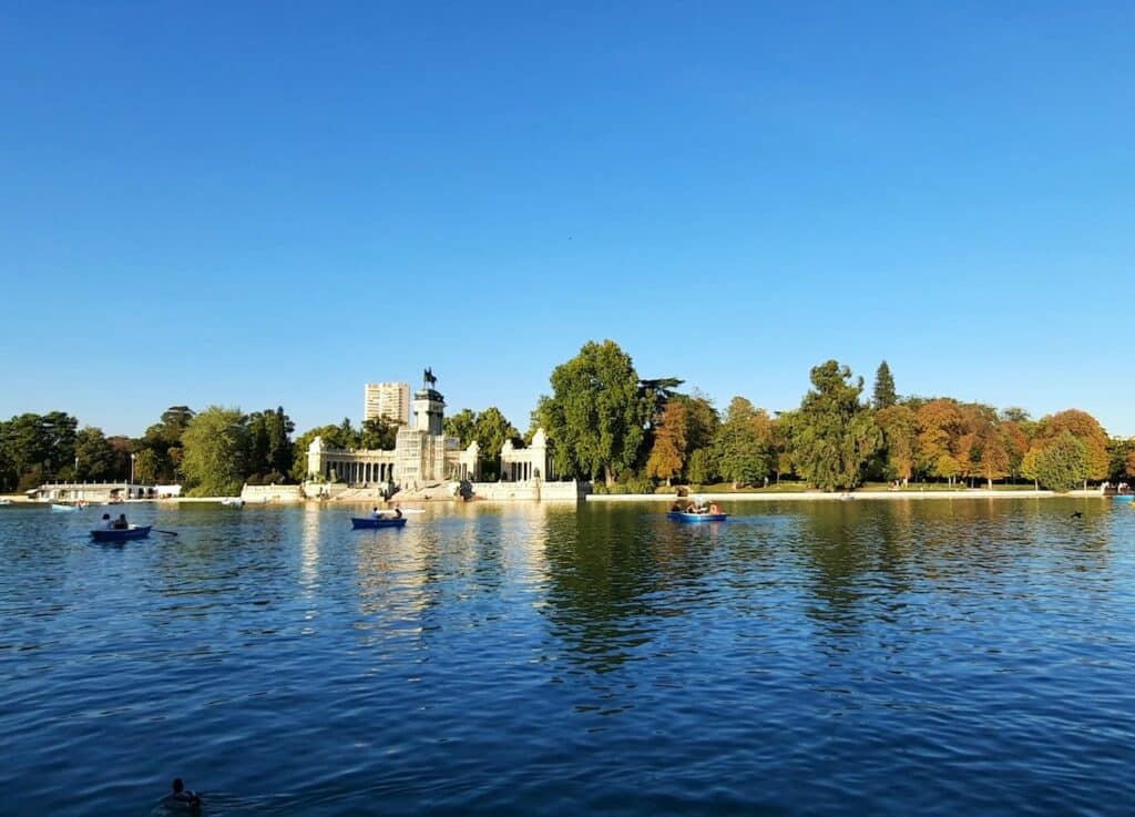 Vista do El Retiro Park, com um lado e pessoas andando de canoa com remos, sendo que ao fundo há árvores e uma construção com estátua, o Monumento a Afonso XII
