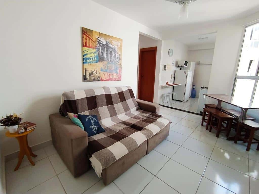 Parte do MAR & SOL Apartamentos I - Free Wi-Fi que mostra a sala com um sofá retrátil, com um quadro em cima na parede, uma mesinha redonda ao lado e em frente uma mesa com seis bancos. Ao fundo tem a parte da cozinha aparecendo com outra mesa na parede, uma geladeira e um micro-ondas e do lado esquerdo tem uma porta de madeira, ilustrando post Pousadas em Rio das Ostras.