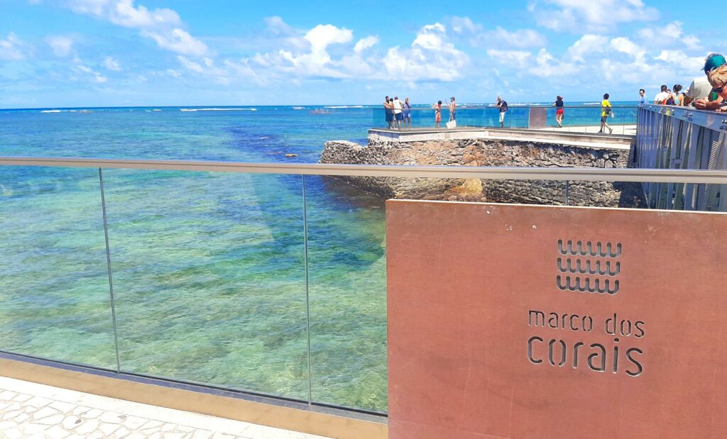 Placa escrito "marco dos corais", com uma praia de água cristalina ao fundo. Há pessoas em volta olhando a paisagem