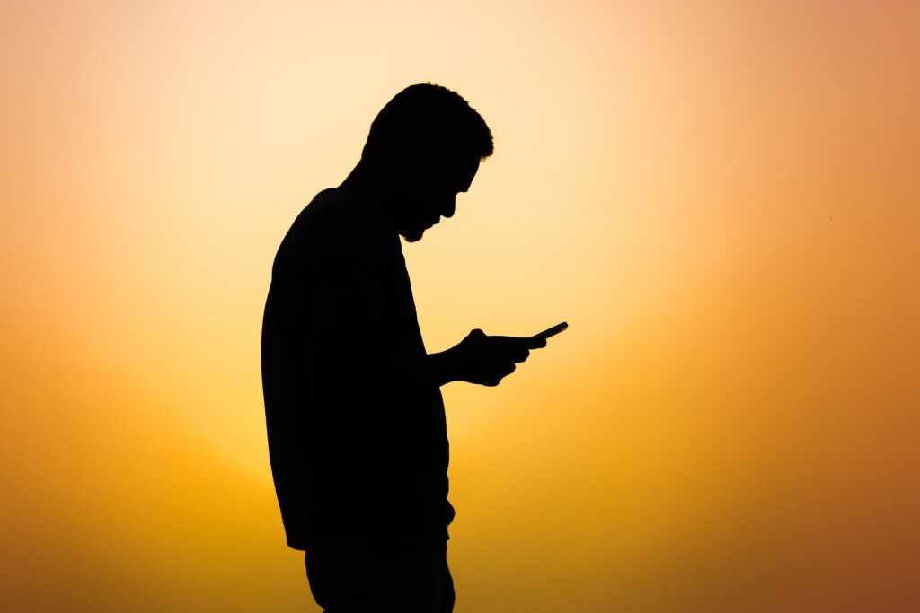 silhueta de um homem de lado, segurando um celular com uma mão. Ao fundo é possível ver uma forte luz amarelada.