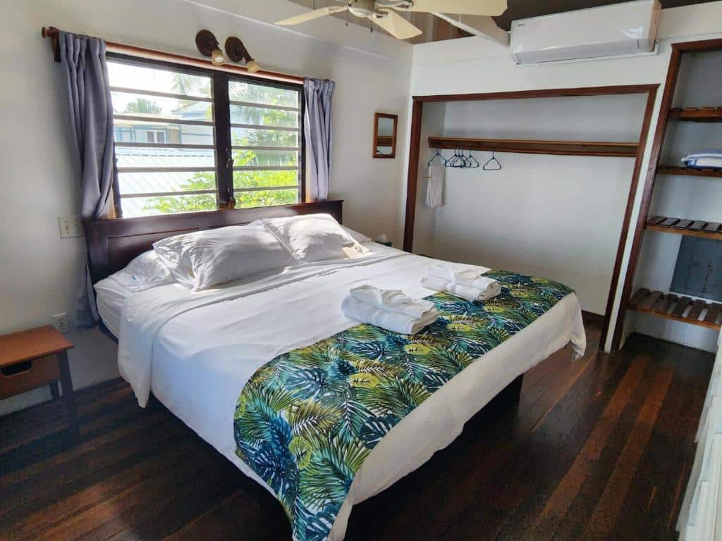 Um quarto no Maya Beach Hotel. A cama está no centro da foto, é box e de casal. Atrás dela há uma janela. Do seu lado esquerdo há uma mesa de cabeceira, tomadas e interruptor. Do seu lado direito há uma arara de roupas com cabides.