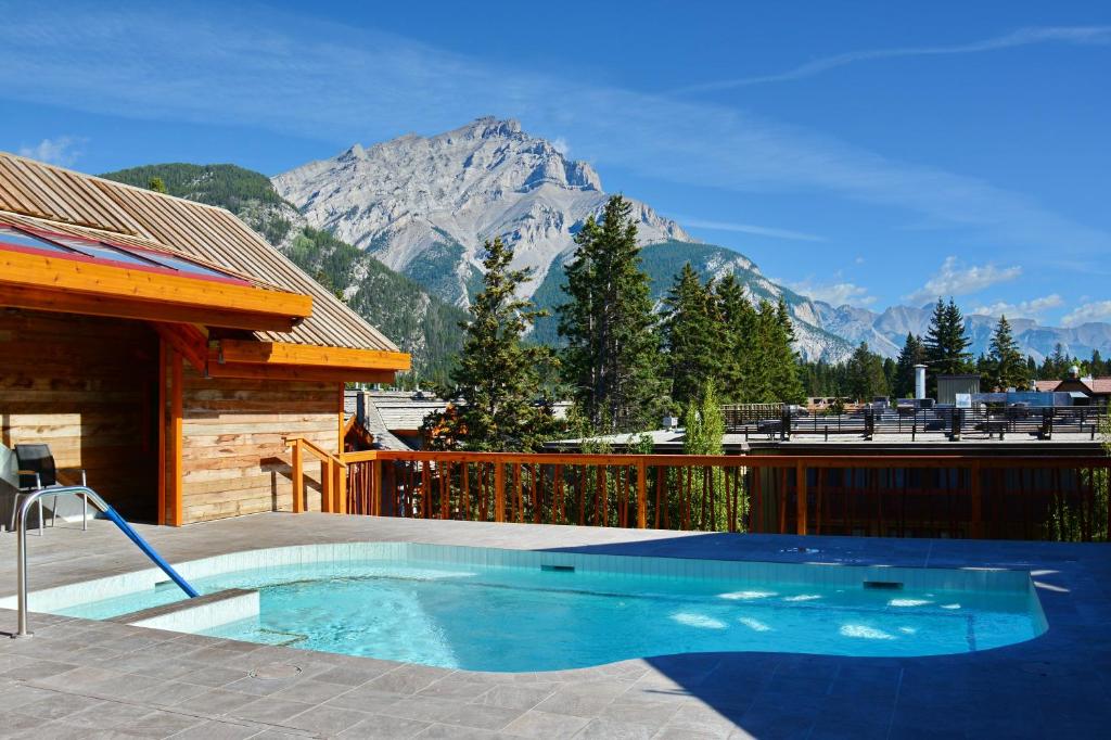 Parte do Moose Hotel and Suites que mostra a piscina ao ar livre, com uma casa de madeira do lado esquerdo e uma grade de madeira em frente a piscina. A vista da piscina dá para algumas construções e árvores e mais ao fundo uma montanha com neve durante o dia, ilustrando post Hotéis em Banff.