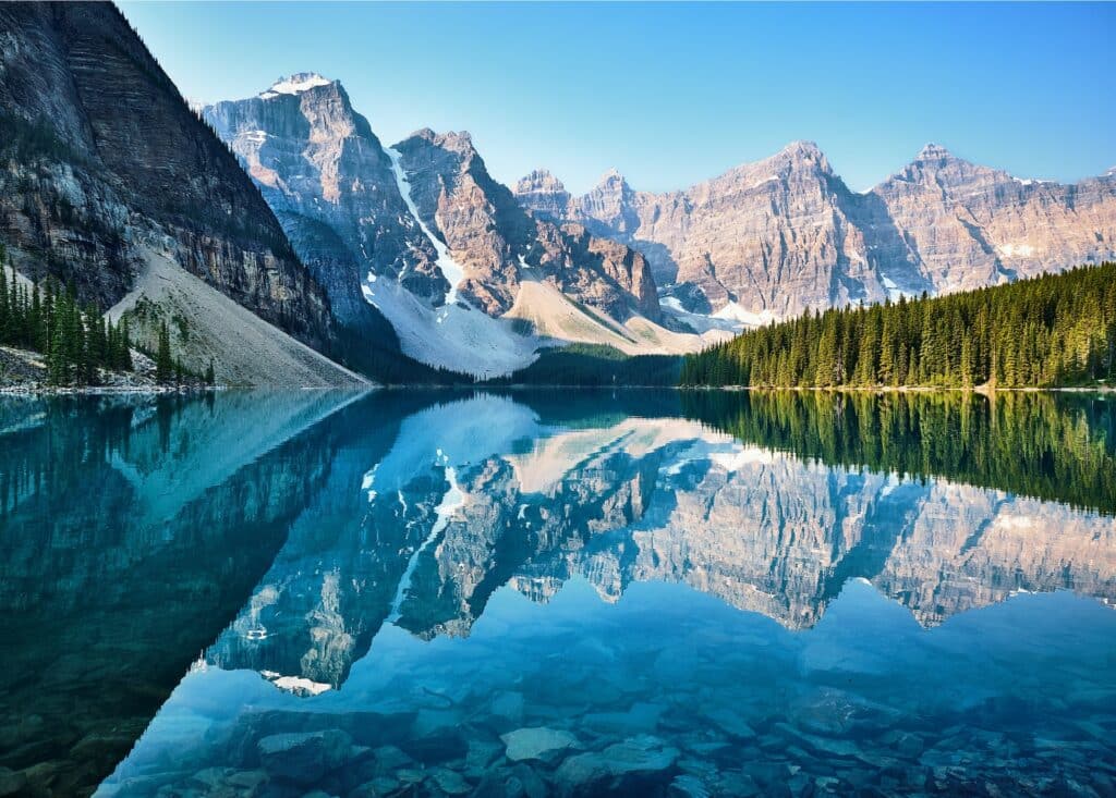 Grande lago no meio de montanhas e floresta para ilustrar o post sobre seguro viagem Canadá. A imagem das montanhas ao fundo está refletida no lado no centro da foto, e a floresta está ao fundo no lado direito. - Foto: John Lee via Unsplash
