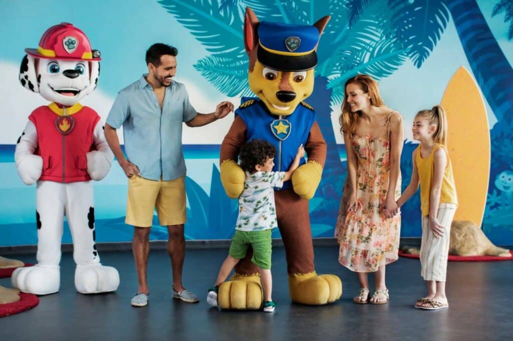 Família conhecendo a Patrulha Canina no Nickelodeon Hotels & Resorts Punta Cana. Da esquerda para a direita: uma pessoa fantasiada de dálmata com roupa de bombeiro; um homem sorrindo enquanto vê um menino abraçando uma pessoa fantasiada de cachorro com uniforme de policial; uma mulher de vestido segura a mão de uma menina, e as duas sorriem para o garoto.