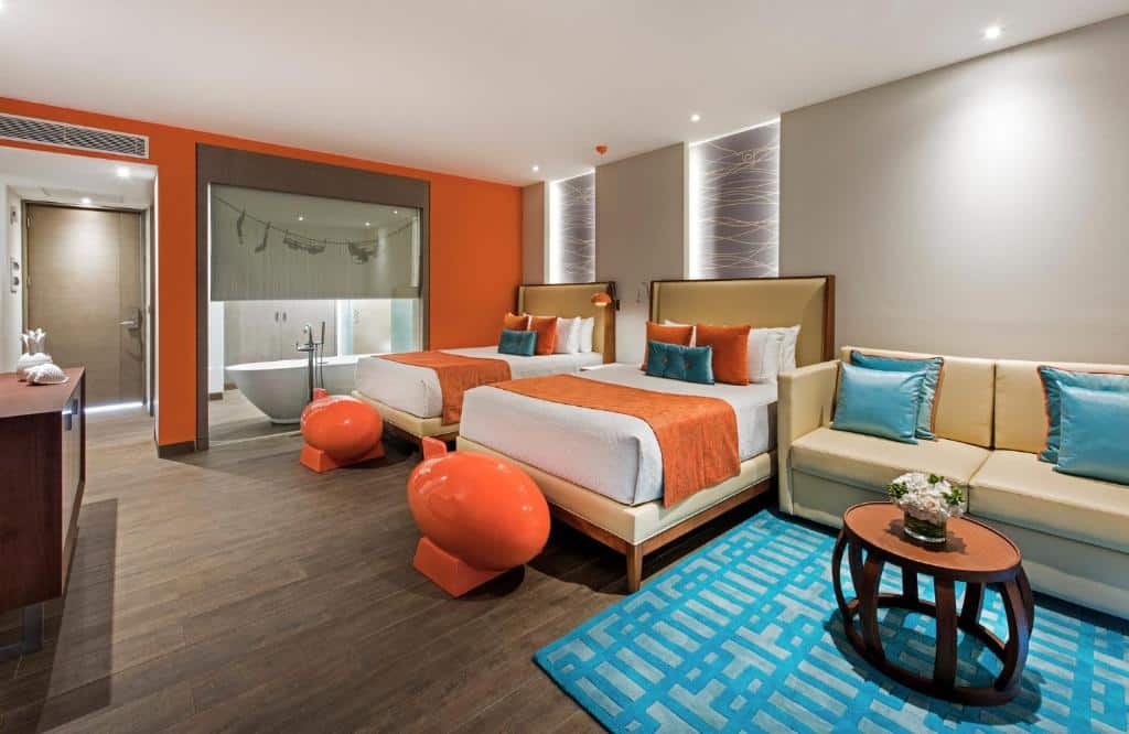 Quarto do Nickelodeon Hotels & Resorts Punta Cana, um dos resorts all inclusive em Punta Cana. No canto direito fica uma área de estar dom sofá, mesa de centro e tapete. Ao lado esquerdo há duas camas de casal separadas por uma mesinha de cabeceira e uma luminária. Estátuas de um foguete laranja estão aos pés das duas camas, que encaram uma cômoda na parede oposta. Ao fundo e do lado esquerdo de uma das camas é possível ver uma banheira no banheiro.