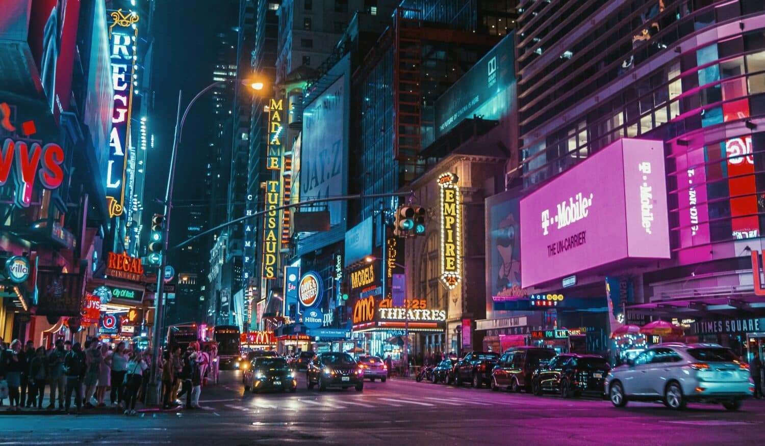 avenida da Times Square vista de noite, com vários painéis iluminados e coloridos margeando as ruas com diversos carros e pessoas em movimento.
