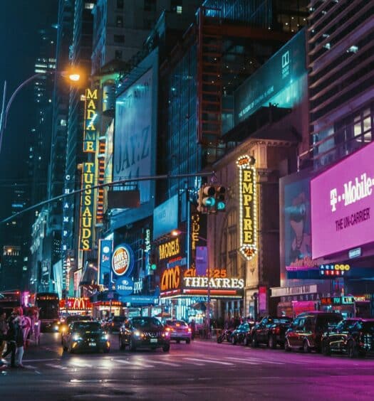 avenida da Times Square vista de noite, com vários painéis iluminados e coloridos margeando as ruas com diversos carros e pessoas em movimento.