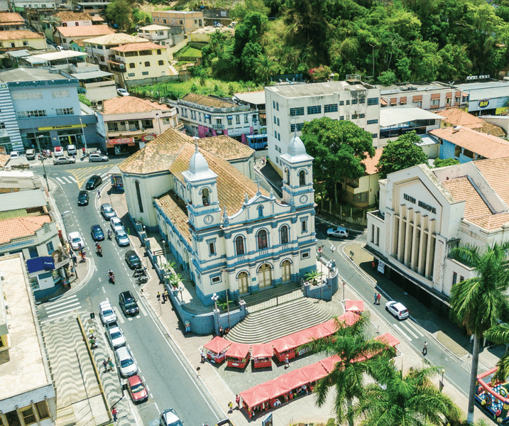Vista aérea da Igreja Matriz de Nova Lima. Ao redor vemos outras lojas, casas, construções, árvores, e carros e motos passando na rua. O Teatro Municipal está do lado direito da igreja.