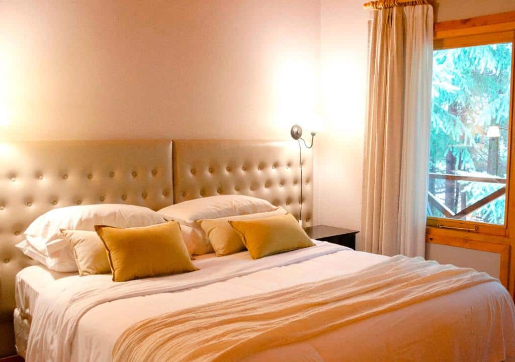 Uma cama de casal no ONA Apart Hotel and Spa, com uma cabeceira estofada. No canto direito a janela do quarto. Foto para ilustrar post de hotéis em Villa La Angostura.