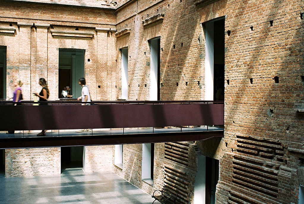 Museu da Pinacoteca de São Paulo. Uma ponte no meio com barras de apoio, ao redor paredes com tijolinho e diversas janelas. Em baixo, um banquinho de madeira.