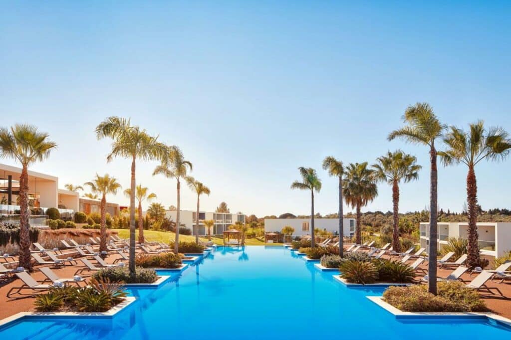 Piscina do Tivoli Alvor Algarve – All Inclusive Resort no centro da imagem, em cada lado da piscina coqueiros e cadeiras.