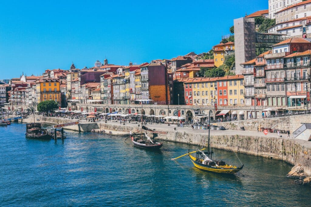 Vista do Cais da Ribeira no Porto, com barcos no rio Douro e ao fundo casinhas coloridas.